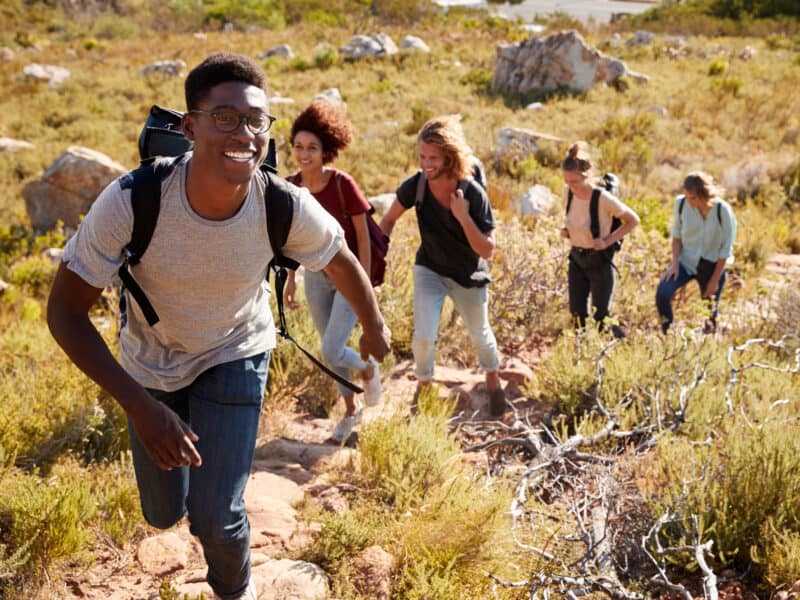 Millennial African American man leading friends hiking single file uphill on a path in countryside