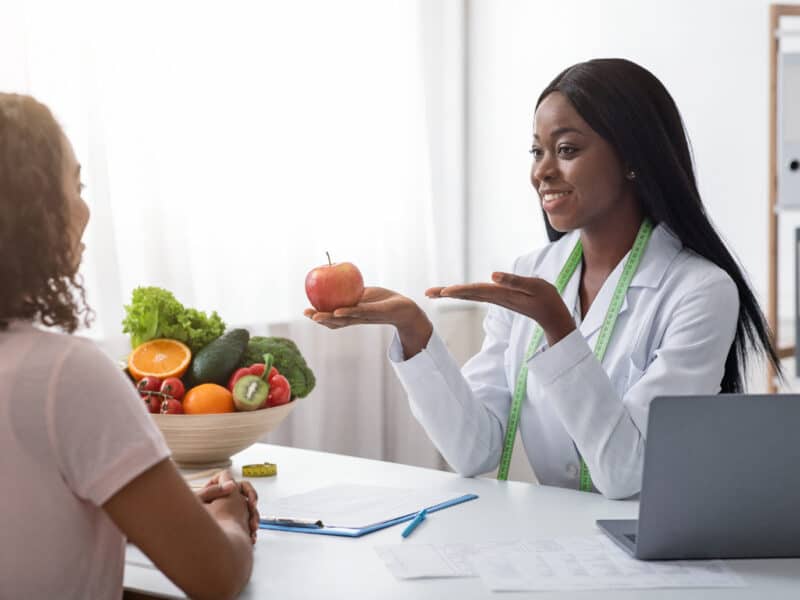 Positive african woman nutritionist recommending female patient fresh fruits to eat, clinic interior